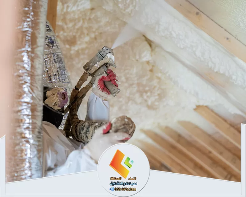 عامل يرتدي ملابس حماية يرش عزل فوم أبيض على سقف خشبي مائل داخل مبنى باستخدام مسدس رش مخصص.